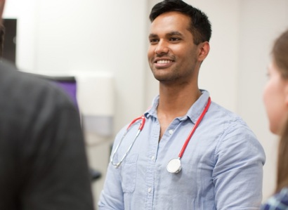 A photo of a young doctor with a stethoscope around his neck. He is looking to the left of the image, where 2 other people can be seen out of focus, with a woman on the right-hand side of the image also visible.