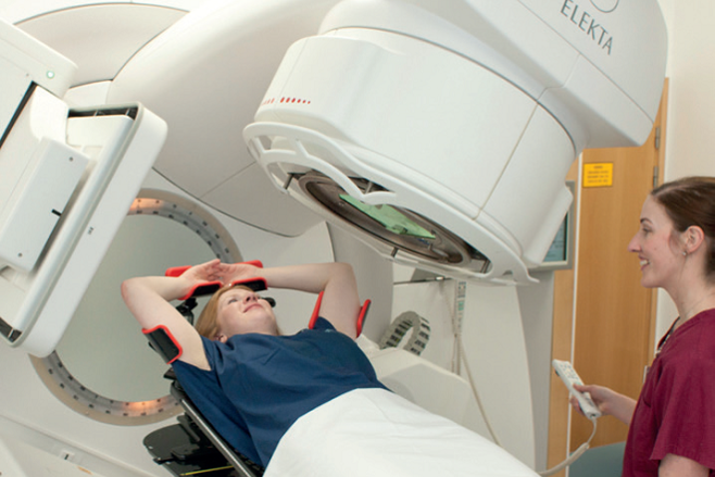 A photo of a patient lying on their back on the radiotherapy treatment machine called a linear accelerator (or linac). The patient has their arms above their head placed in arm rests. To the side of the patient is a radiographer. The treatment machine arm extends from the linac and is positioned above and to the side of the patient. There is an imaging board on the other side of the patient.