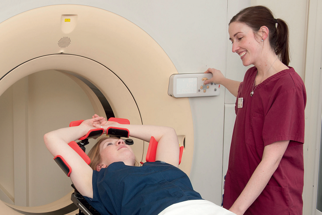 An image of a patient lying on their back on the CT scanner bed. The patient has their arms above their head placed in arm rests and the CT scanner can be seen behind them. To the side of the patient is a radiographer.