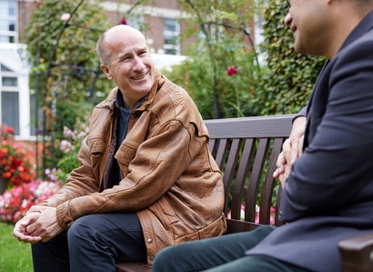 A photo of a male patient sitting on a bench in The Christie garden speaking to a male staff member who is mainly out of shot.