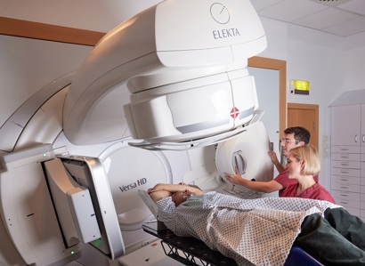 A photo of a male patient receiving radiotherapy on an Elekta Linac machine with a male radiographer and a female radiographer.