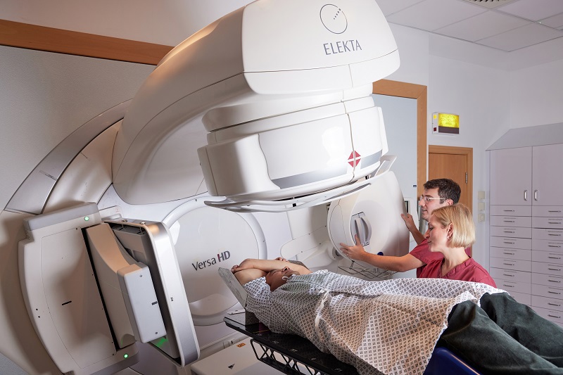 A photo of a male patient receiving radiotherapy on an Elekta Linac machine with a male radiographer and a female radiographer.