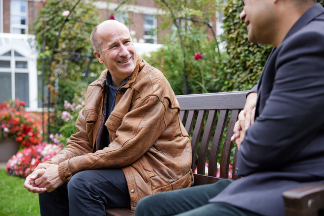 A photo of a male patient sitting on a bench in The Christie garden speaking to a male staff member who is mainly out of shot.