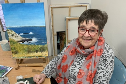 A photo of Christie supporter Linda in the art room at The Christie in front of one of her paintings of a beach scene.