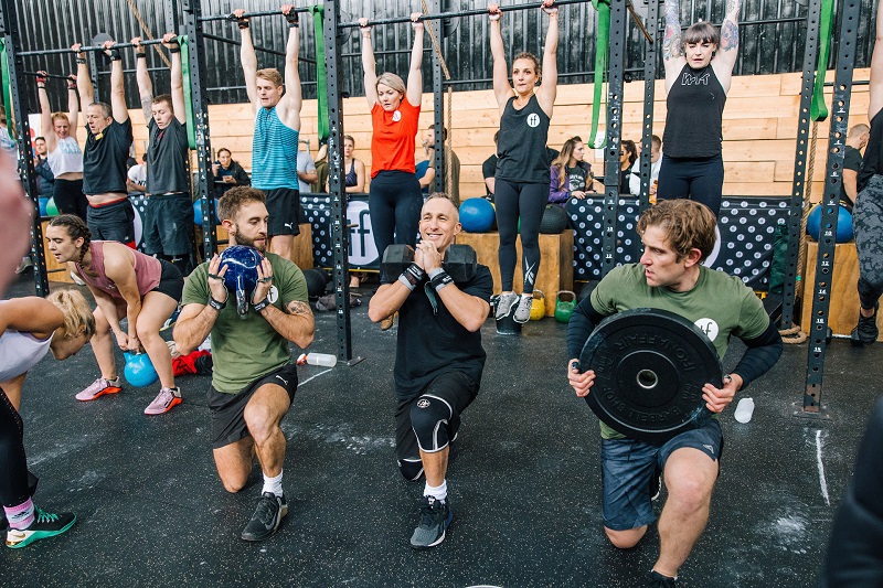 A photo of a group of people working out. In the foreground, 3 men and 1 woman are lifting a range of weights; in the background, 4 women and 3 men are hanging off exercise bars, while several people spectate.