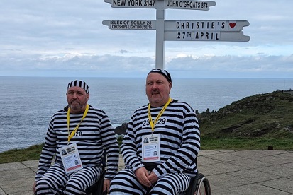 A photo of Christie fundraisers Carl and Peter dressed in black and white striped outfits as convicts and both sitting in wheelchairs. The 2 are in front of the sign for Land's End which reads 'New York 3147', 'Isles of Scilly 28', 'Longships Lighthouse 1 1/2', 'John O'Groats 874' and 'Christie's 27th April'.