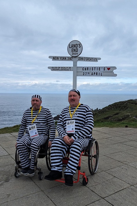 A photo of Christie fundraisers Carl and Peter dressed in black and white striped outfits as convicts and both sitting in wheelchairs. The 2 are in front of the sign for Land's End which reads 'New York 3147', 'Isles of Scilly 28', 'Longships Lighthouse 1 1/2', 'John O'Groats 874' and 'Christie's 27th April'.