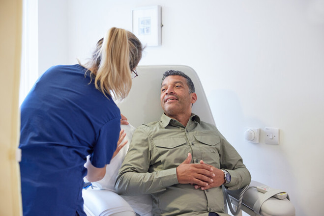 A photo of a man sitting in a hospital treatment chair, speaking to a female staff member.