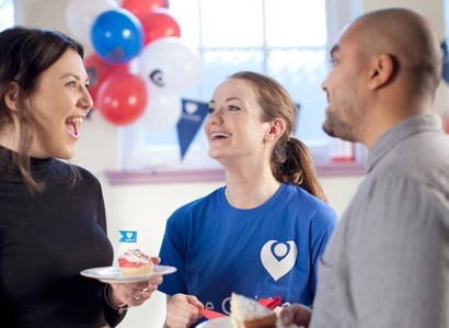 A photo of a fundraising event for The Christie Charity. One female fundraiser in the middle of the photo is wearing a blue Christie Charity t-shirt and she has another female fundraiser on one side and a male fundraiser on the other side. They are holding cake, and balloons and bunting can be seen in the background.