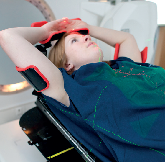 A photo of a patient on their back on the CT scanner bed. The patient has their arms above their head placed in arm rests and the CT scanner can be seen behind them. There are some lights from a laser beam projected on the chest of the patient which help line up the patient in the correct treatment position.