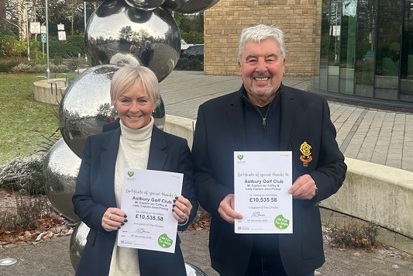 A photo of Astbury Golf Club captains Ian Coffey and Jane Pickup standing outside The Christie at Macclesfield entrance; both are holding certificates thanking Astbury Golf Club for raising £10, 535.58 for The Christie Charity.