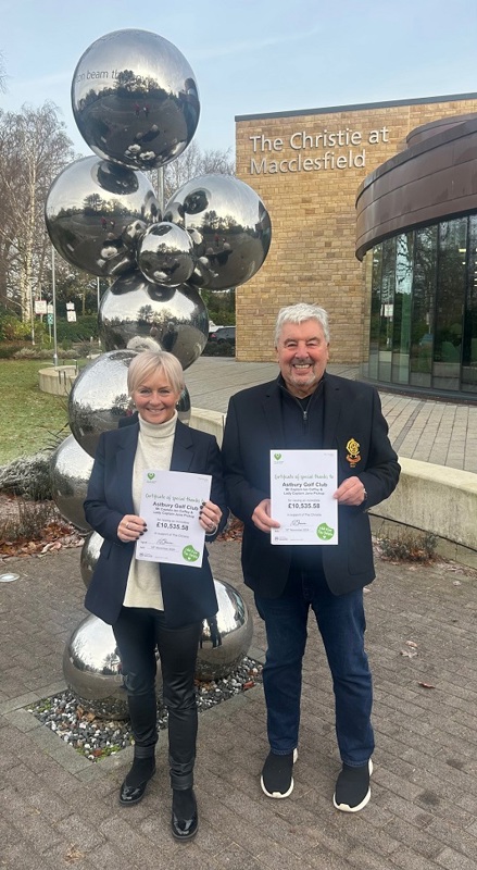 A photo of Astbury Golf Club captains Ian Coffey and Jane Pickup standing outside The Christie at Macclesfield entrance; both are holding certificates thanking Astbury Golf Club for raising £10, 535.58 for The Christie Charity.