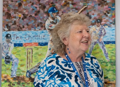 A photo of Pat Mountford, the artist in residence at The Christie, standing in front of a painting by a Christie patient.