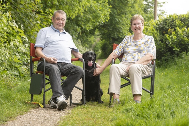 A photo of Christie patient Eileen Unsworth sitting in a chair on the right with her husband Peter on the left and their black Labrador Annie sitting between them.