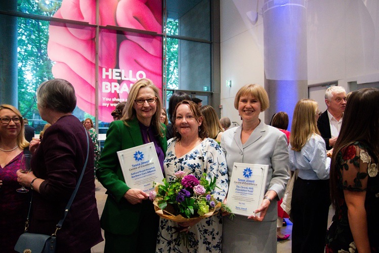 A photo of Tina Karabatsou, another woman and Catherine McBain. Tina and Catherine are holding plaques reading 'Tessa Jowell Centre of Excellence, Salford Royal Hospital, Northern Care Alliance', and the middle woman is holding a bunch of flowers.